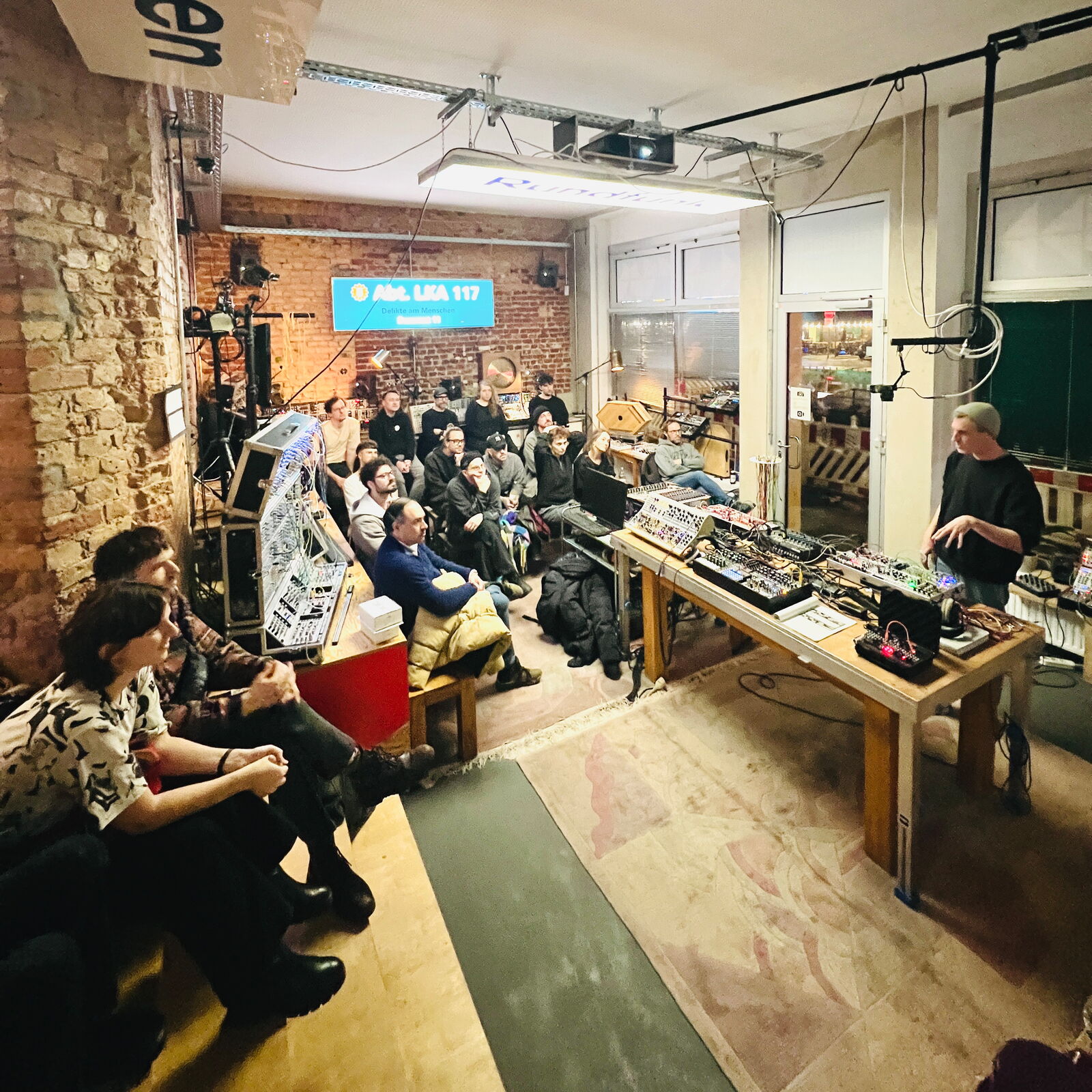 Workshop evening at SchneidersLaden in Berlin: a large audience listens as the presenter explains the basics at a table full of modular synthesizers