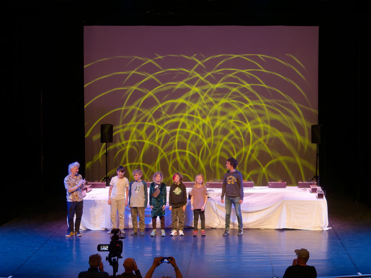 Children from the Modular Synthesizer Ensemble on stage after their performance, with synthesizers and a light projection behind them as the workshop leader applauds